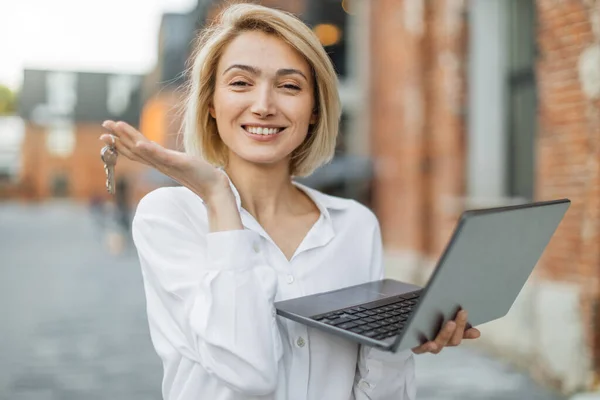 Real estate attractive agent beautiful woman with house keys and laptop in front of sold apartment, in city street. Young caucasian female in white shirt with keys just bought new apartment.