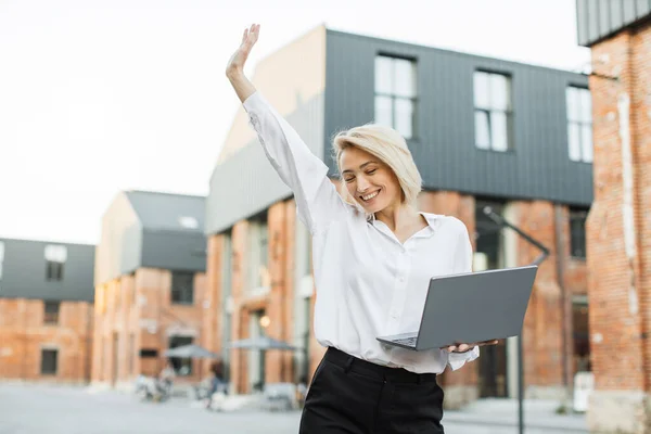 Happy satisfied caucasian business woman, relaxing while walking on near office building raises hand up while rejoicing with laptop doing online order after shopping. Success and happy news concept.