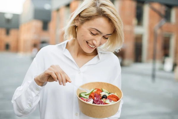 街の建物を背景にサラダを食べる若い魅力的な女性の肖像画 新鮮なサラダを屋外で楽しむ美しい若い女性 可愛いですブロンド女の子食べるA健康的な食事 — ストック写真