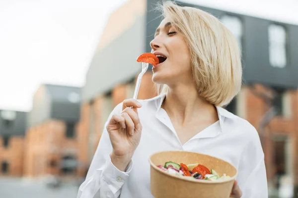 一方で新鮮なサラダのボウルを保持し 他方でフォークにトマトのスライスをかむ肯定的な空腹ブロンドの女性の肖像画 健康的な食事のコンセプト — ストック写真