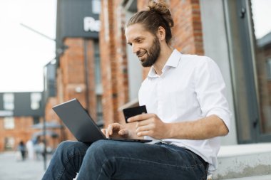 İnternetten alışveriş yapmak için laptop ve kredi kartı kullanan beyaz gömlekli yakışıklı bir adam. Teneffüste merdivenlerde oturan sakallı iş adamı..