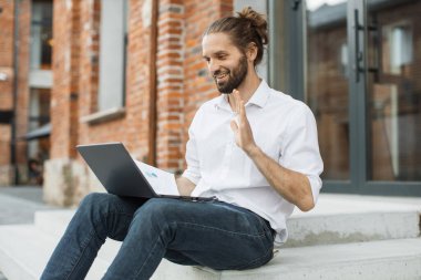 Gülümseyen mutlu adam dışarıda çalışırken bilgisayarında video konferansı yapıyor. Çekici beyaz erkek merdivenlerde oturuyor ve grafik ve çizelge gösteriyor..