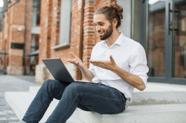Gülümseyen mutlu adam dışarıda çalışırken bilgisayarında video konferansı yapıyor. Çekici beyaz erkek merdivenlerde oturuyor ve grafik ve çizelge gösteriyor..