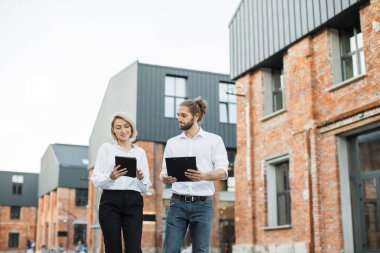 İki Kafkasyalı ofis çalışanı dışarıda birlikte yürüyüp gülümsüyor ve sohbet ediyorlar. Beyaz gömlekli yakışıklı bir adam, elinde pano ve elinde tablet tutan çekici bir kadın..