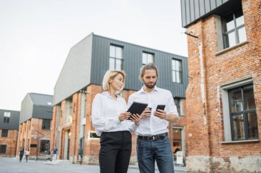 Dijital tabletli çekici bir kadın ve akıllı telefonlu çekici bir adam ve ofis binasının yakınında birlikte duran bir fincan kahve. İki meslektaş dışarıda çalışmak için modern aletler kullanıyor..