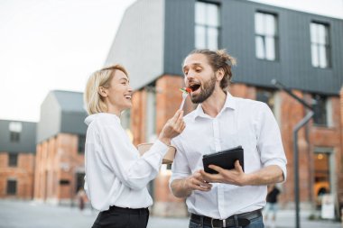 Dışarıda gülerek ve sohbet ederek yürüyen iki mutlu ofis çalışanı. Çekici bir kadın sağlıklı salatayla kase taşıyor ve erkek arkadaşını besliyor, elinde tablet var..