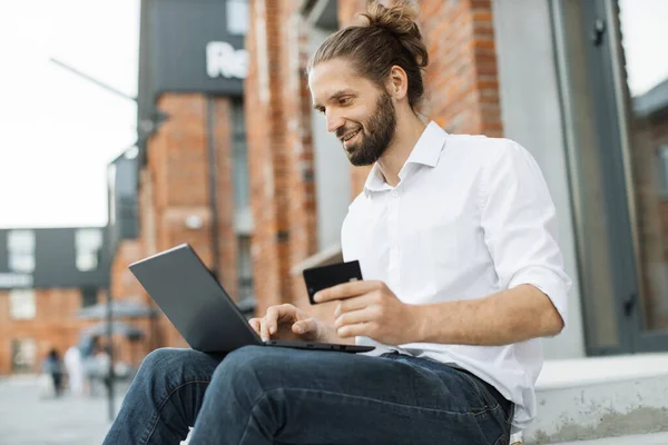 Handsome attractive man in white shirt using laptop and credit bank card for online shopping. Bearded businessman sitting on stairs outdoor during break and making purchase.