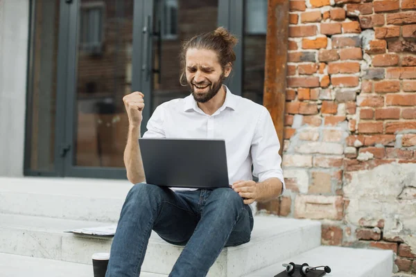 Happy satisfied caucasian businessman, relaxing while sitting on the steps of a office building rejoicing with laptop doing online order after shopping. Success and happy news concept.
