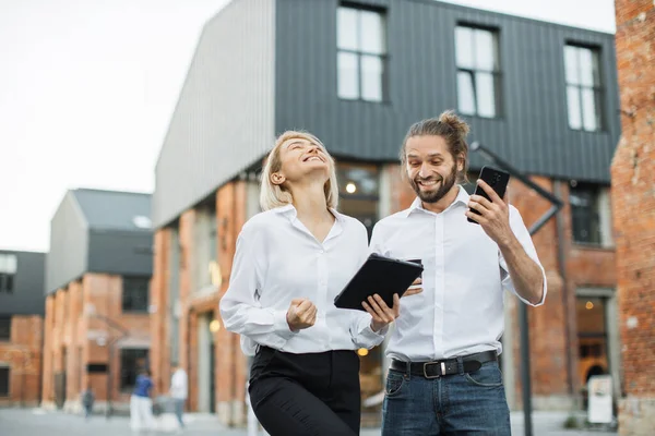 Two happy excited office workers walking together outdoors smiling and chatting. Handsome man carrying smartphone and cup of coffee, charming woman holding tablet.