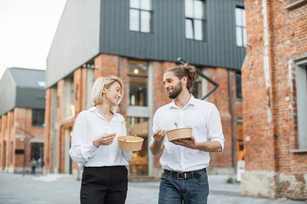 Öğle yemeğinde sağlıklı salata yiyen mutlu gençler. Neşeli iş ortakları şehir sokaklarında yürümeye ara veriyor ve birlikte gülen yeşil sebzelerle vejetaryen yemeği yiyorlar..