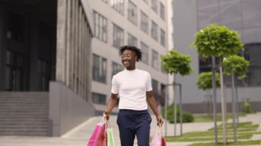 Şık giyinmiş neşeli Afrikalı bir kızın şehirde alışveriş yaparken çekilmiş yatay bir fotoğrafı. Modern ofis binalarının arka planında alışveriş torbalarıyla yürüyen Afro-Amerikalı bir kadın.