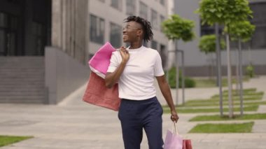 Genç, mutlu, gülümseyen, Afro-Amerikan bir kadın, elinde kese kağıtları, modern şehir binalarının caddelerinde yürüyor. Kopyala, metin için boşluk.
