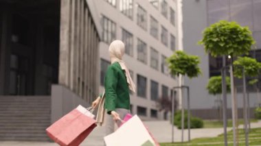 Açık havada, başörtüsü ve gözlük takan, şehirde yürüyüş ve alışverişin keyfini çıkaran, gülümseyen ve renkli alışveriş çantalarıyla dolaşan zarif, modaya uygun Müslüman kadının şehir portresi..