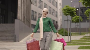 Alışveriş, satış, satın alma konsepti. Genç, modaya uygun Müslüman bir kadın elinde renkli alışveriş poşetleri, kolları havada sokakta poz veriyor. Arka planda bulanık modern şehir.