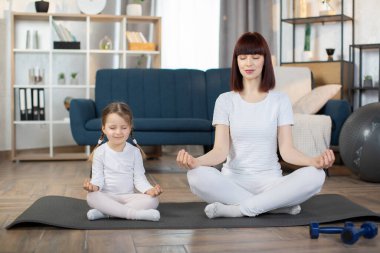 Güzel genç bir kadın ve sevimli küçük kızı evde yoga yaparken gülümsüyorlar. Anne ve tatlı küçük kız, gözleri kapalı otururken yoga minderinde poz verirken gülümsüyor.