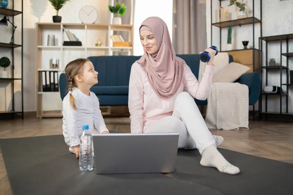 Young muslim mom and daughter doing sports, fitness exercises with laptop on the floor at home. Sporty arabic mother with child girl exercising in the morning. Healthy family lifestyle concept.