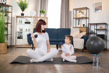 Spor, spor konsepti. Anne ve Küçük Kız evdeki oturma odasındaki yoga minderinde dambıl egzersizi yapıyorlar. Yeni normal konsept. Sağlıklı bir yaşam tarzı. Evde dinleniyorum..