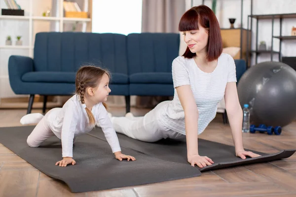 A young caucasian mom with her little daughter doing fitness, stretching, push up exercises on the floor mat in the living room. Sports at home and female beauty.