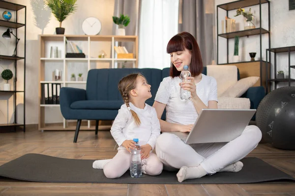 Mother and daughter engaged in yoga in sportswear sitting on mat at home in a bright room drinking water from the bottles while watching online fitness video course on a laptop.
