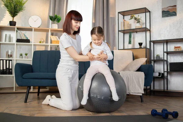 Happy fitness family training on swiss ball in living room. Portrait of sporty positive caucasian mother enjoying home fitness activity with her little cute girl sitting on rubber fitball.