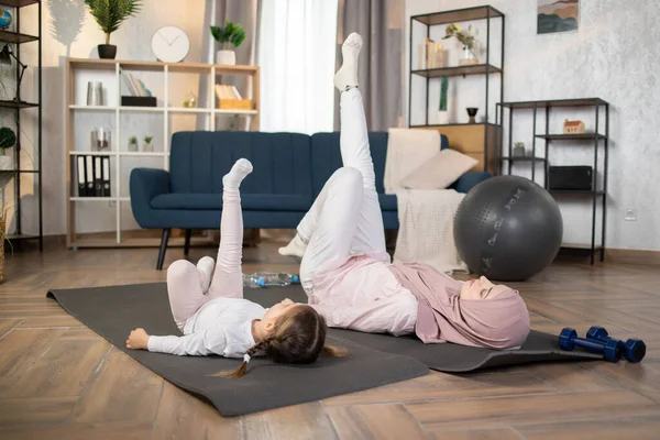 Young positive muslim woman doing stretching flexibility exercises together with her preschool daughter, happy family practicing gymnastics at home, fitness training with child.
