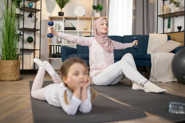 Attractive positive happy muslim mom doing sports using dumbbells while her cute little girl is resting lying on the mat in living room at home.