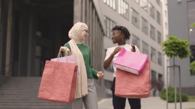 Açık havada, şık giyinmiş, kameraya poz veren ve modern şehirde renkli alışveriş torbaları taşıyan iki genç bayanın moda portresi..