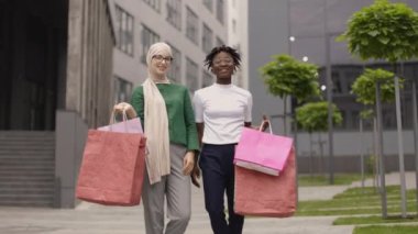Şehir alışveriş konsepti. Modern şehirde yürürken ellerinde renkli alışveriş torbalarıyla gezen iki farklı Afrikalı ve Müslüman kadının şehir dışı yaşam tarzı portresi..