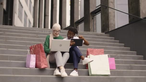 两个快乐而又满足的多民族女孩 坐在现代化建筑的台阶上 一边放松 一边在网上购物 一边享受着网上购物的乐趣 在城市购物 城市生活方式 — 图库视频影像
