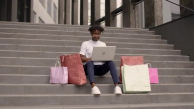 Açık havada, şık giyinmiş Afrikalı genç bir kadının moda portresi büyük bir alışveriş merkezinin merdivenlerinde oturuyor ve online renkli alışveriş torbaları sipariş ediyor..