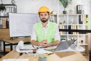 Sarı kasklı ve yansıtıcı yelekli sakallı mühendis büyük televizyon ekranının arka planında robot resimle birlikte masada oturuyor. İş adamı ortak inşaat projesi üzerinde çalışıyor