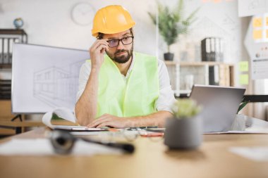 Yansıtıcı yelek, baret ve gözlüklü uzman mimar masanın üzerinde dizüstü bilgisayar ve plan ile inşaat projesini kontrol ediyor. Sakallı çekici adam çalışma sürecini kontrol ediyor..