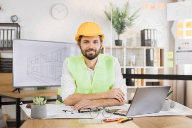 Uzman mimar erkek, masa başında dijital dizüstü bilgisayarla oturuyor ve pusulayı proje çizimleri için kullanıyor. Üniformalı, yeşil yansıtıcı yelek giyen sakallı genç adam ve inşaat başlığı..