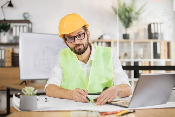 Architect man or male builder hold pen and ruler, writing and drawing architectural project, work on laptop computer on the desk at workplace looking at camera.