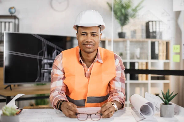 迷人的非洲工程师头戴白色头盔 身穿反光背心 坐在桌旁 背景上有大量的蓝图和草图 从事建筑工程的商人 — 图库照片