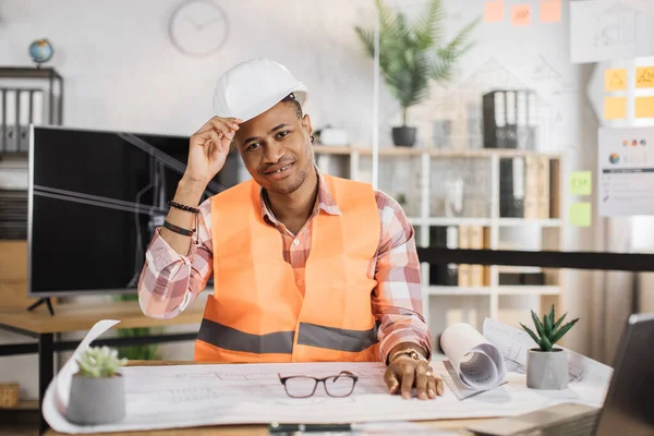 Attractive African engineer in white helmet and reflective vest sitting at table with lots of blueprints on the background of large tv screen with sketch. Business man working on construction project.