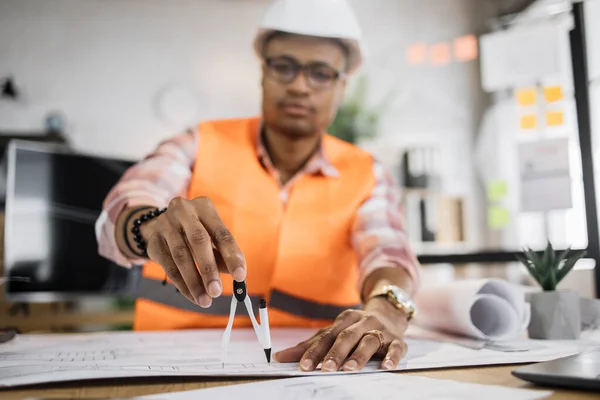 Focus on compass. Young african builder, engineer in reflective vest uniform and hard hat on desk in office on background of large TV screen with .