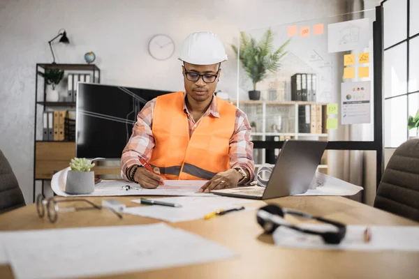 African American architect man or male builder hold pen and ruler, writing and drawing architectural project, work on laptop computer on the desk at workplace.