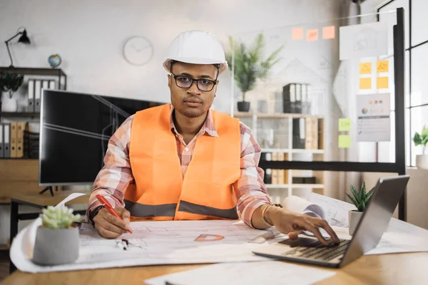 Portrait of african american man wearing uniform and white hard hat at work. Confident contractor sitting at the desk, holding pen and working on laptop with sketches of building object.