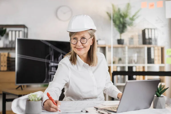 Mature architect woman or female builder holding pen while writing and drawing architectural project using laptop computer, sitting on the desk at workplace in modern office.