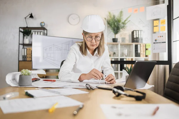 Competent mature female engineer, designer or architect leading working conference at office center. Attractive young senior woman wearing protective helmet working with compass, laptop and blueprint.