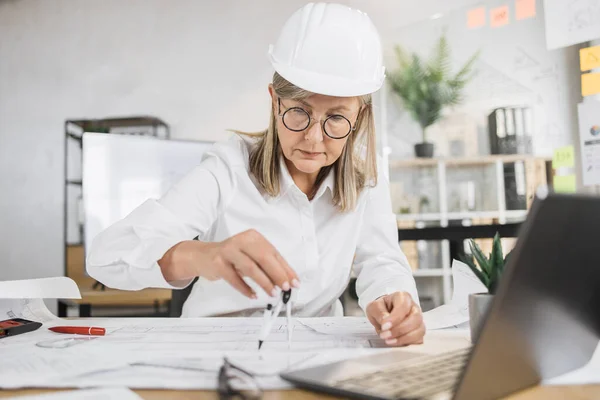 Competent mature female engineer, designer or architect leading working conference at office center. Attractive young senior woman wearing protective helmet working with compass, laptop and blueprint.