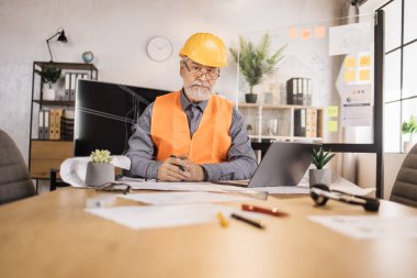 Yetişkin mühendis, tasarımcı ya da mimar ofis merkezinin önde gelen iş konferansı. Üniformalı ve koruyucu başlıklı çekici yaşlı sakallı adam dizüstü bilgisayarında planlarla çalışıyor..