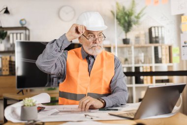Beyaz koruyucu kasklı ve üniformalı profesyonel kıdemli erkek mimar yeni dijital dizüstü bilgisayar kullanıyor, inşaat planı üzerinde çalışıyor, iş yerinde çizimler yapıyor..