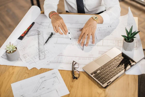 Close up of hands of architect, engineer or designer sitting at table and analysing blueprints sitting at desk in office. Creation of construction project.