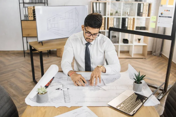 Qualified arab architect in white shirt, tie and glasses checking construction project with blueprint and laptop sitting on table. Bearded attractive muslim man controlling working process.