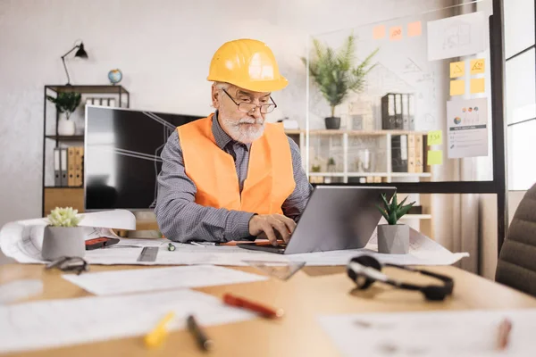 Bearded engineer senior man in helmet and reflective vest sitting at table with lots of blueprints on the background of large tv screen with sketch. Business man working on common construction project