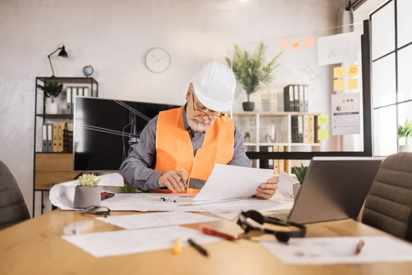 Senior builder, engineer in reflective vest uniform and hard hat on desk in office on background of large TV screen with , browsing in laptop and looking through papers or sketches.