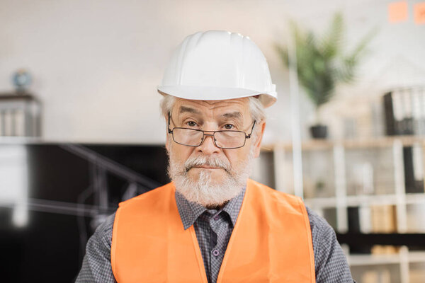 Portrait of mature architect wearing white construction helmet indoors. Bearded man in orange reflective vest posing among architectural office of the company.
