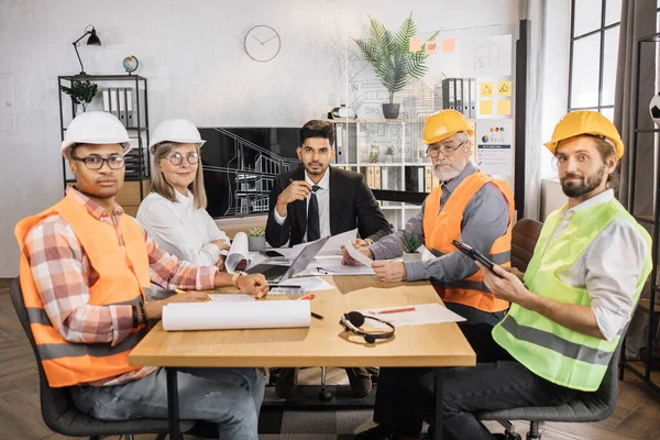 Qualified architects, engineers and designers having business meeting at office in front of TV screen. Multicultural colleagues in suits and helmets sitting at table and looking at camera.